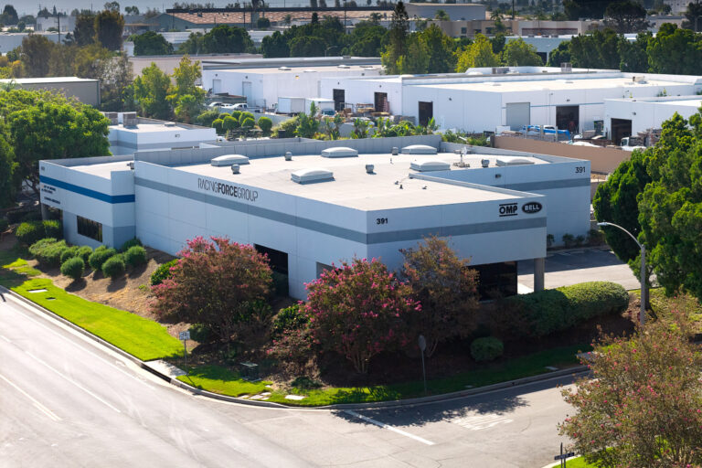 A professional, realistic corporate and industrial photo inside the modern Racing Force Group West Coast Operations Center in Corona, California. Clean, well-lit showroom and technical workshop floor displaying high-tech OMP racing suits, Bell motorsport carbon-fiber helmets, and precision racing equipment on sleek minimalist shelving. Through glass partitions, warehouse logistics racks and clean office spaces are visible. Bright sunny California daylight coming through windows, sharp focus, architectural and commercial photojournalism, 8k resolution.